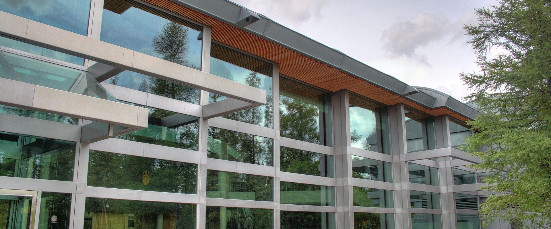 A modern building with a glass facade, metal framing, and wooden ceiling accents under an overhang, with picnic tables outside.