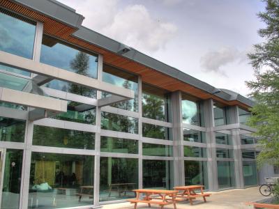 A modern building with a glass facade, metal framing, and wooden ceiling accents under an overhang, with picnic tables outside.