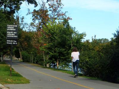 A paved pathway alongside a body of water with a person walking away from the camera, featuring a bilingual sign prohibiting commercial vehicles and a speed limit sign.