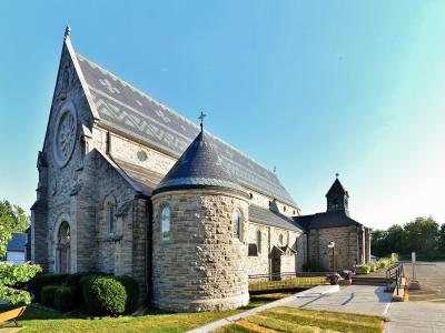 A stone Roman Catholic church with a prominent apse, bell tower, and slate roof, set against a blue sky with a paved walkway and parking area in the foreground.