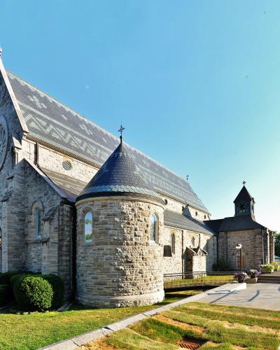 A stone Roman Catholic church with a prominent apse, bell tower, and slate roof, set against a blue sky with a paved walkway and parking area in the foreground.