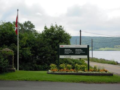 A roadside sign for the Alexander Graham Bell National Historic Site of Canada, featuring bilingual text and the Parks Canada logo, situated next to a flagpole with a Canadian flag and a body of water.