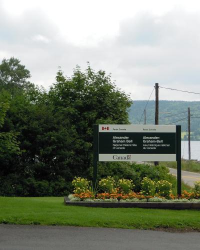 A roadside sign for the Alexander Graham Bell National Historic Site of Canada, featuring bilingual text and the Parks Canada logo, situated next to a flagpole with a Canadian flag and a body of water.