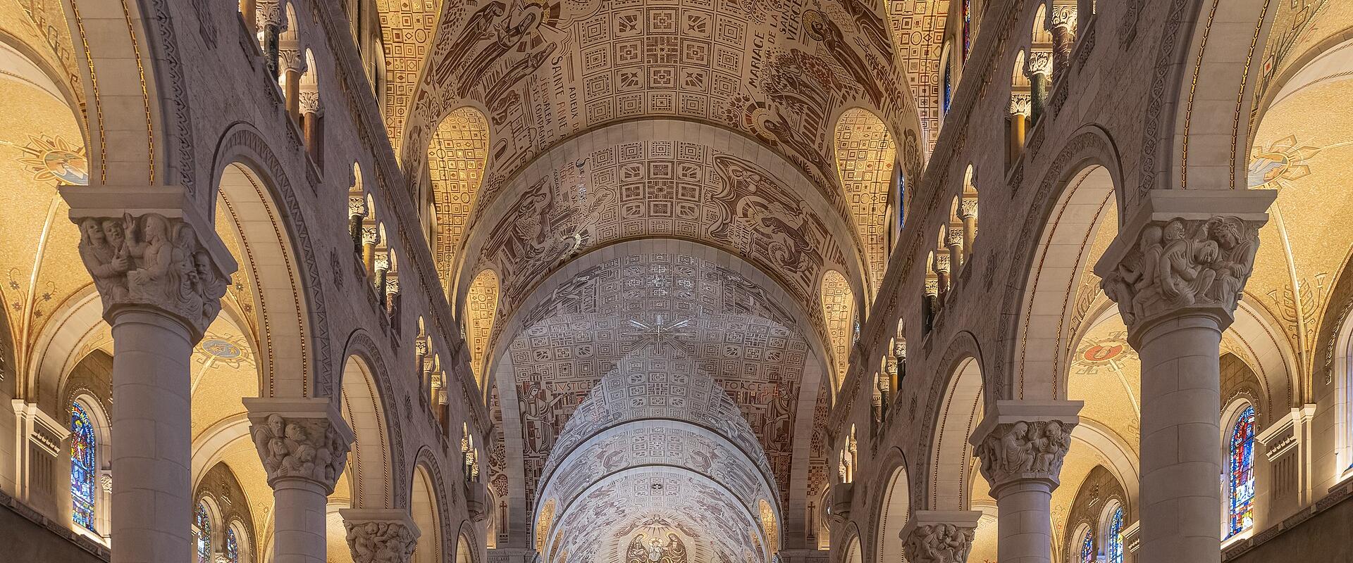 The image shows the interior nave of a large Romanesque Revival basilica, featuring a highly decorated barrel-vaulted ceiling with extensive mosaics, rows of wooden pews, and large stone columns.