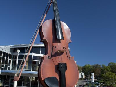 A large, reddish-brown sculpture of a fiddle with a bow, standing on a paved plaza in front of a modern building with large glass windows under a clear blue sky.