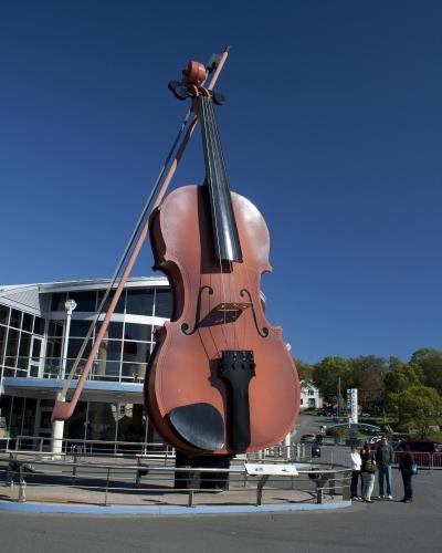 A large, reddish-brown sculpture of a fiddle with a bow, standing on a paved plaza in front of a modern building with large glass windows under a clear blue sky.