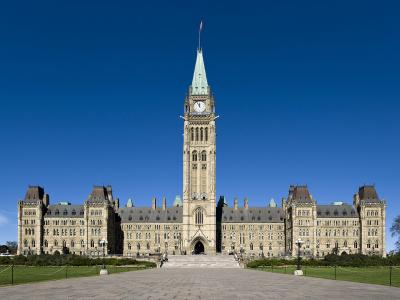 A low-angle, wide shot of a large neo-Gothic stone government building with a prominent central clock tower topped by a Canadian flag, set against a clear blue sky.