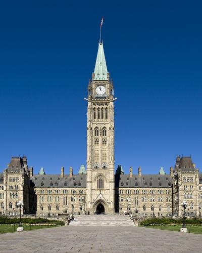 A low-angle, wide shot of a large neo-Gothic stone government building with a prominent central clock tower topped by a Canadian flag, set against a clear blue sky.