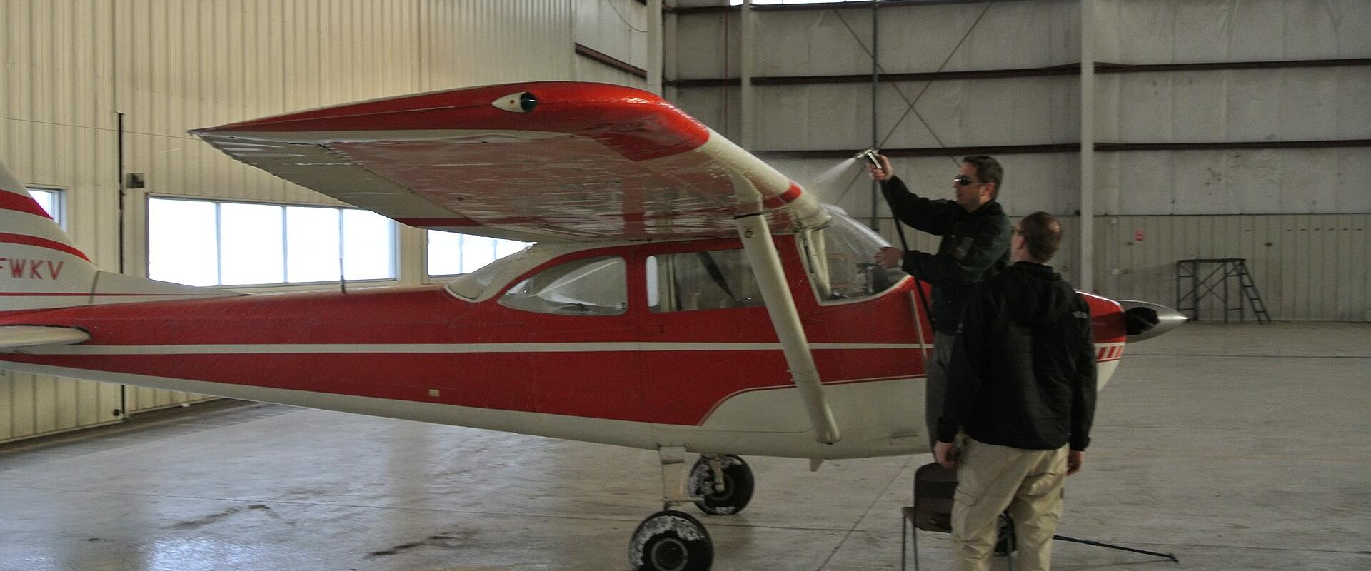 A red and white single-engine aircraft is inside a hangar while a person sprays its wing with a hose, with the tail registration C-FWKV visible.