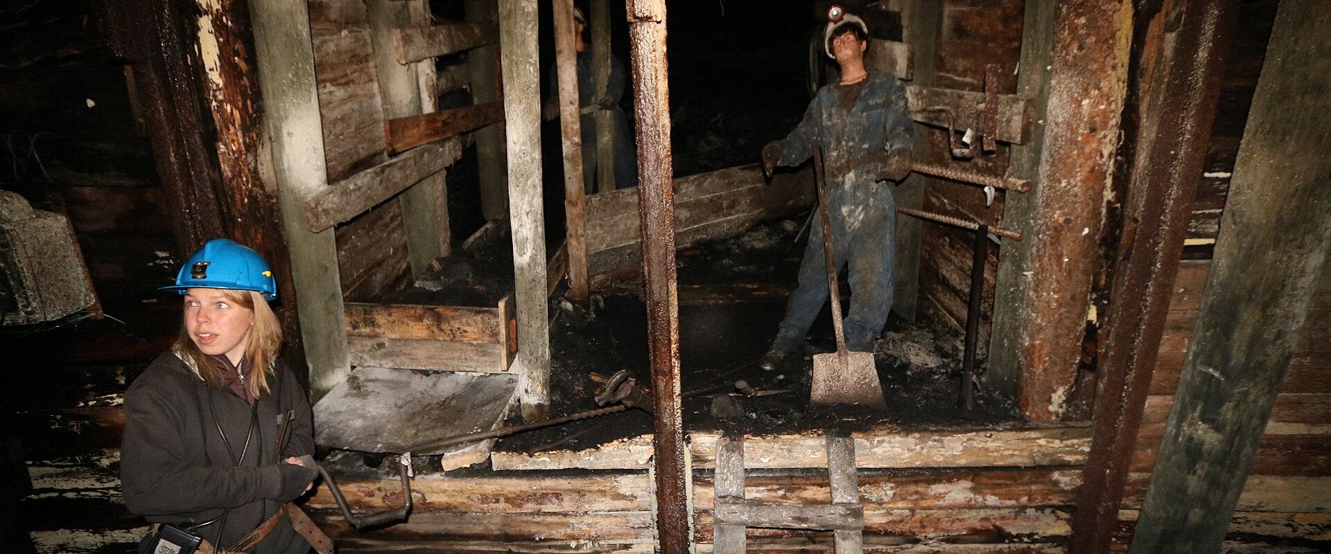 A woman wearing a blue hard hat and carrying a lantern stands in the foreground of a dark, timber-supported underground mine shaft, with a mannequin of a miner holding a shovel visible in the background.