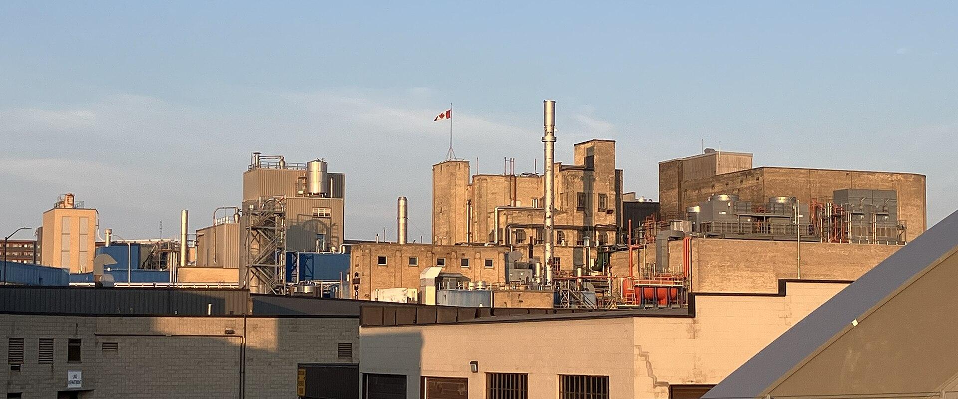 An industrial complex featuring large brick buildings, metal silos, and smokestacks, viewed from a bridge with a concrete railing in the foreground and a Canadian flag flying on a pole.