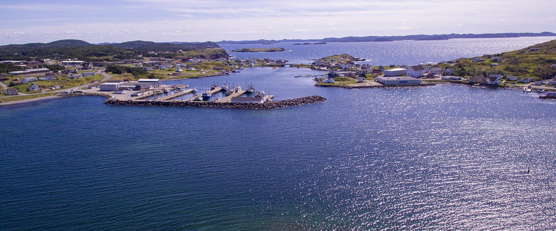 An aerial view of a coastal fishing harbour with a breakwater, several docked fishing vessels, and a small town settlement on the surrounding headlands under a blue sky.