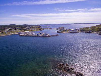 An aerial view of a coastal fishing harbour with a breakwater, several docked fishing vessels, and a small town settlement on the surrounding headlands under a blue sky.