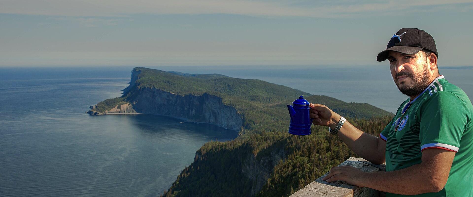 A man in a green shirt and black cap stands at a wooden viewpoint holding a blue coffee pot, overlooking a large, forested peninsula with steep cliffs dropping into the ocean.