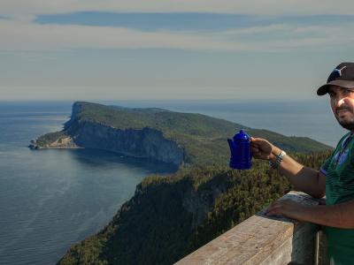 A man in a green shirt and black cap stands at a wooden viewpoint holding a blue coffee pot, overlooking a large, forested peninsula with steep cliffs dropping into the ocean.