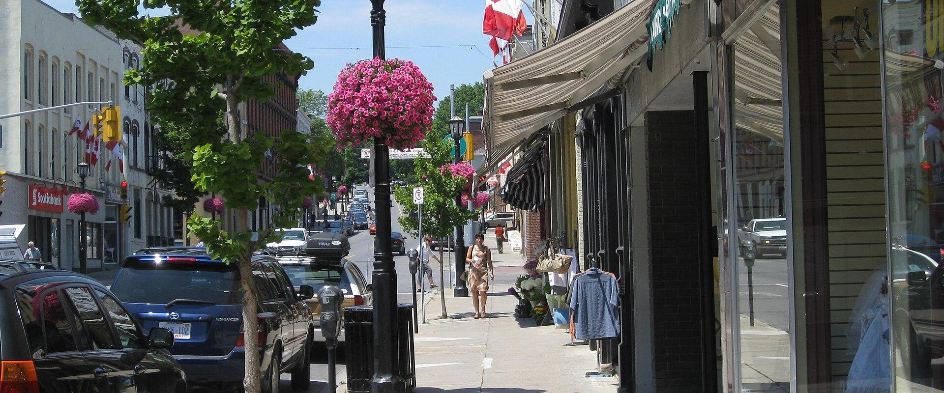 A street-level view of a commercial downtown area with brick buildings, storefronts, parked cars, and Canadian flags hanging from poles along the sidewalk.