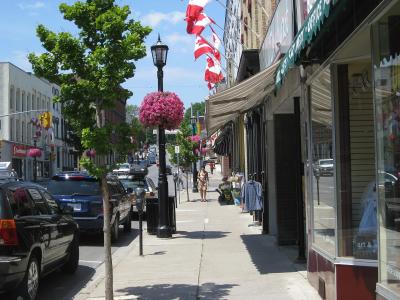 A street-level view of a commercial downtown area with brick buildings, storefronts, parked cars, and Canadian flags hanging from poles along the sidewalk.