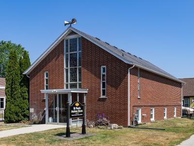 A red brick church building with a tall stained-glass window on the gable end, a weather vane on the roof, and a black sign in the front yard reading "St Paul's Anglican Church".