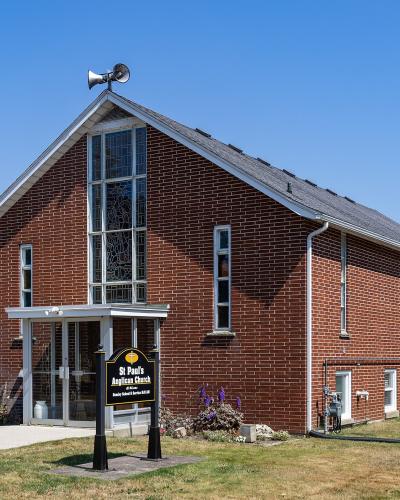 A red brick church building with a tall stained-glass window on the gable end, a weather vane on the roof, and a black sign in the front yard reading "St Paul's Anglican Church".