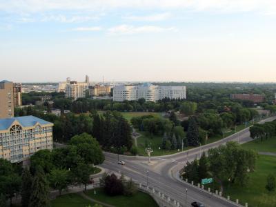 A high-angle view of a city street intersection with a large white hospital building in the background, surrounded by green trees and other urban structures.