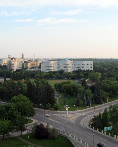 A high-angle view of a city street intersection with a large white hospital building in the background, surrounded by green trees and other urban structures.