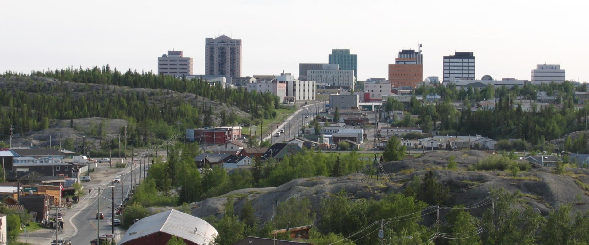 A cityscape with mid-rise buildings rises above rocky hills and green trees, with roads and smaller structures in the foreground under a bright sky.