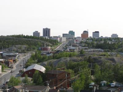 A cityscape with mid-rise buildings rises above rocky hills and green trees, with roads and smaller structures in the foreground under a bright sky.