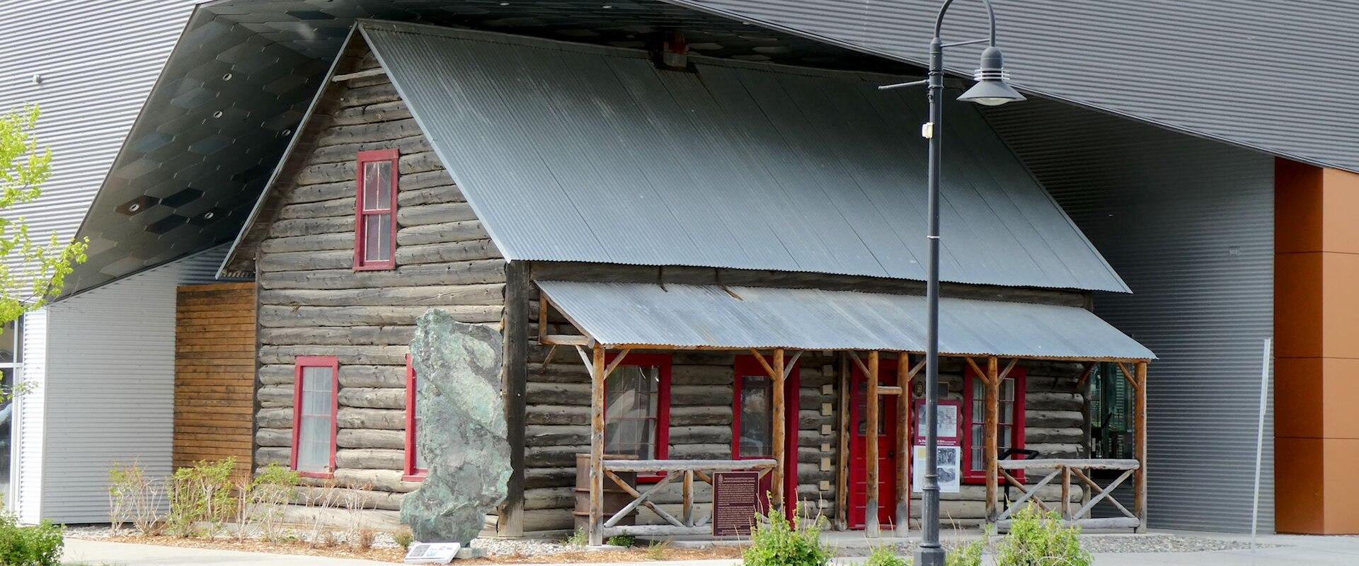 A historic log cabin building with red window frames and a corrugated metal roof, situated directly in front of a large modern building with grey corrugated metal siding.