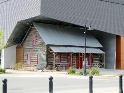A historic log cabin building with red window frames and a corrugated metal roof, situated directly in front of a large modern building with grey corrugated metal siding.