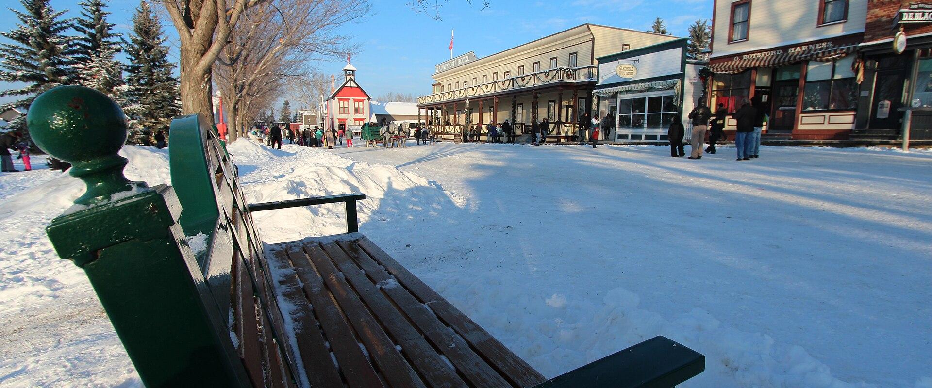 A green wooden bench in the foreground overlooks a snow-covered street lined with historic-style buildings, including a red schoolhouse and a building with a "Ford Harness" sign, with people walking in the distance.