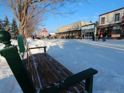 A green wooden bench in the foreground overlooks a snow-covered street lined with historic-style buildings, including a red schoolhouse and a building with a "Ford Harness" sign, with people walking in the distance.