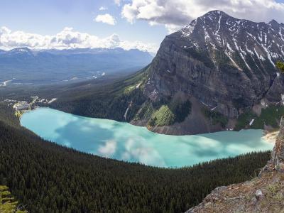 A high-angle panoramic view of a large, turquoise alpine lake surrounded by dense coniferous forests and steep mountain slopes, with a prominent glacier-covered peak rising on the right side.