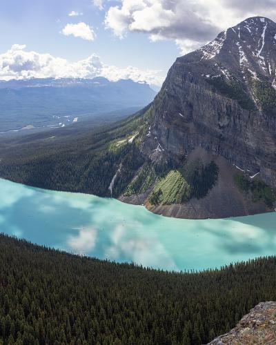 A high-angle panoramic view of a large, turquoise alpine lake surrounded by dense coniferous forests and steep mountain slopes, with a prominent glacier-covered peak rising on the right side.