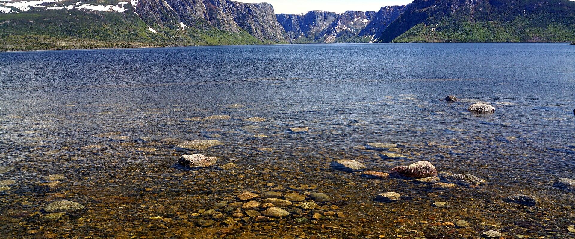 The sheer rock cliffs of the Western Brook Pond fjord in Gros Morne National Park rise above calm blue water, with snow patches on the plateau and rounded boulders visible through the clear shallows in the foreground.