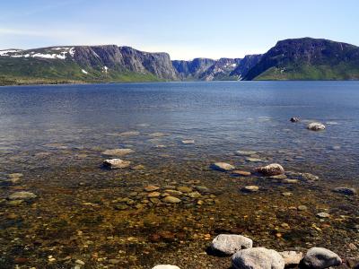 The sheer rock cliffs of the Western Brook Pond fjord in Gros Morne National Park rise above calm blue water, with snow patches on the plateau and rounded boulders visible through the clear shallows in the foreground.