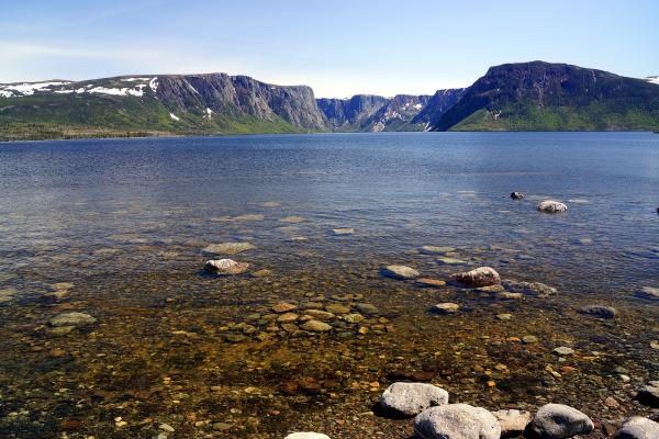 The sheer rock cliffs of the Western Brook Pond fjord in Gros Morne National Park rise above calm blue water, with snow patches on the plateau and rounded boulders visible through the clear shallows in the foreground.