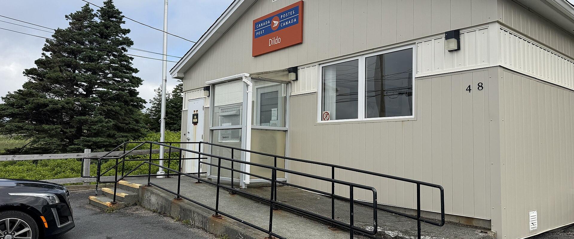 A beige, single-story Canada Post building with a ramp and railing, featuring a sign that reads "CANADA POST / POSTES CANADA" and "Dildo" below it.