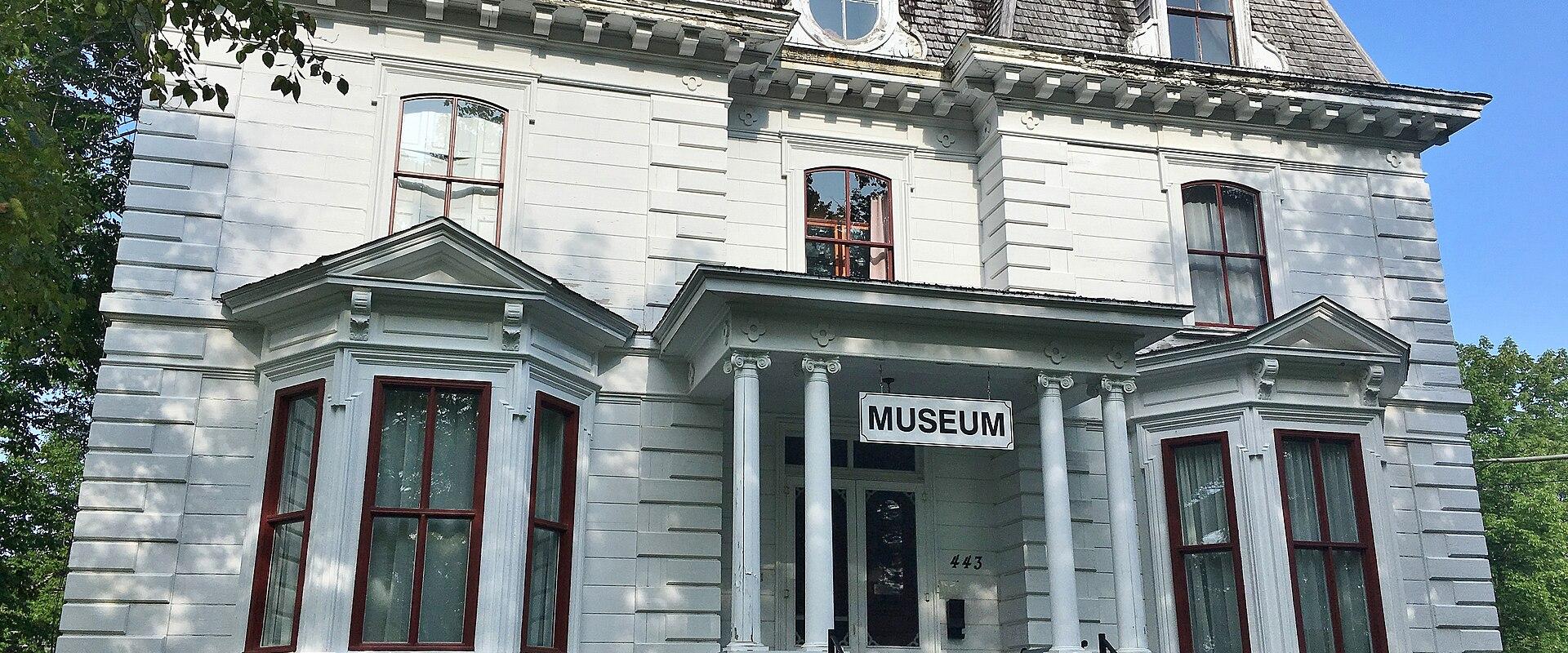 A large, white, three-story Victorian-style house with a mansard roof, red-trimmed windows, and patriotic bunting, featuring a prominent "MUSEUM" sign above the front porch entrance.