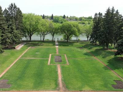 A high-angle view of a formal garden with geometric grass plots and brick pathways leading toward a river, with the Delta Bessborough Hotel visible in the background.