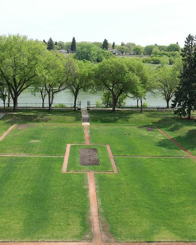 A high-angle view of a formal garden with geometric grass plots and brick pathways leading toward a river, with the Delta Bessborough Hotel visible in the background.