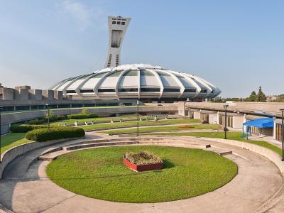 A panoramic view of the Montreal Olympic Park, featuring the distinctive inclined tower and the circular stadium roof in the background, with a curved concrete amphitheater and manicured lawns in the foreground.