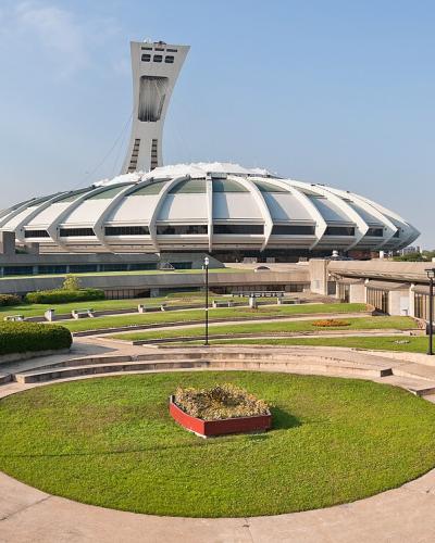 A panoramic view of the Montreal Olympic Park, featuring the distinctive inclined tower and the circular stadium roof in the background, with a curved concrete amphitheater and manicured lawns in the foreground.