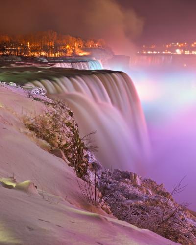 A long-exposure night photograph of a massive waterfall cascading over a cliff edge, with snow-covered ground in the foreground and illuminated mist rising from the base.