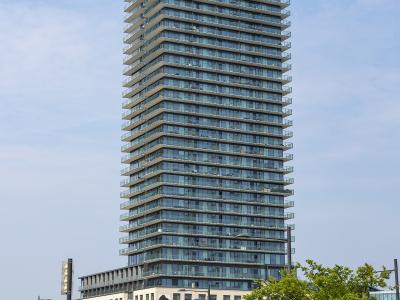 A tall, modern glass-and-concrete residential tower with a stepped design, situated at a street intersection with a red bus and street signs visible at the base.