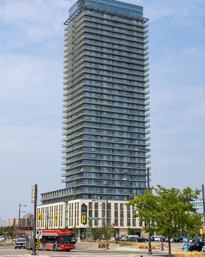 A tall, modern glass-and-concrete residential tower with a stepped design, situated at a street intersection with a red bus and street signs visible at the base.