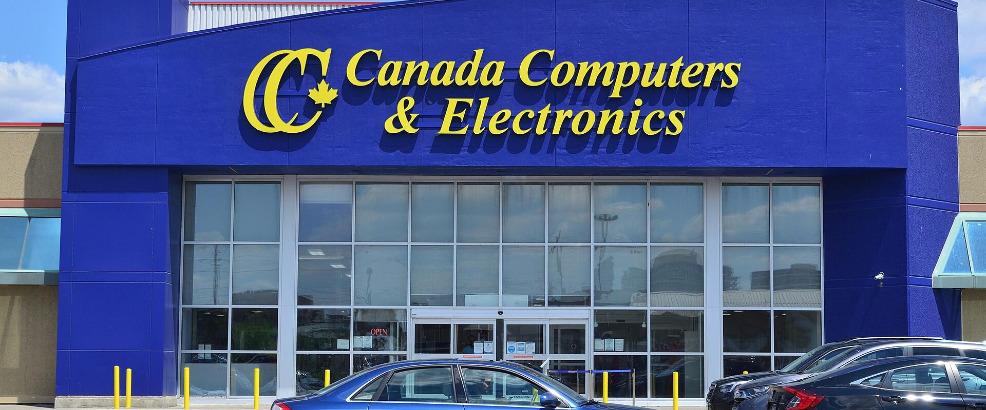 A blue commercial building with a large yellow "Canada Computers & Electronics" sign above glass storefront windows, with cars parked in the foreground.