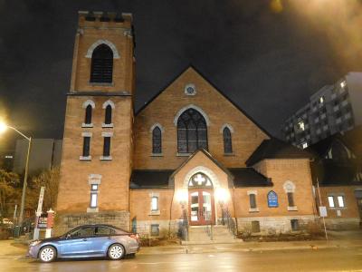A night photograph of a brick Anglican church with a prominent square tower on the left, arched stained-glass windows, and a blue heritage plaque mounted on the wall to the right of the entrance.