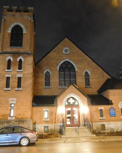 A night photograph of a brick Anglican church with a prominent square tower on the left, arched stained-glass windows, and a blue heritage plaque mounted on the wall to the right of the entrance.