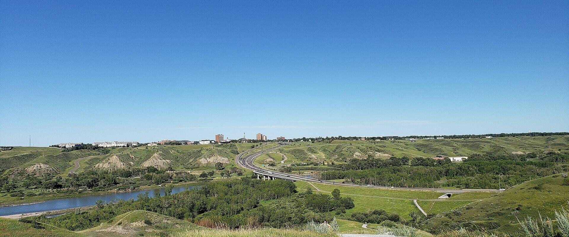 A gravel path leads through rolling green hills toward a river, with a highway and distant city skyline under a clear blue sky.