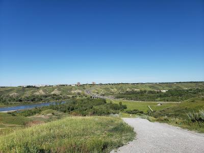 A gravel path leads through rolling green hills toward a river, with a highway and distant city skyline under a clear blue sky.
